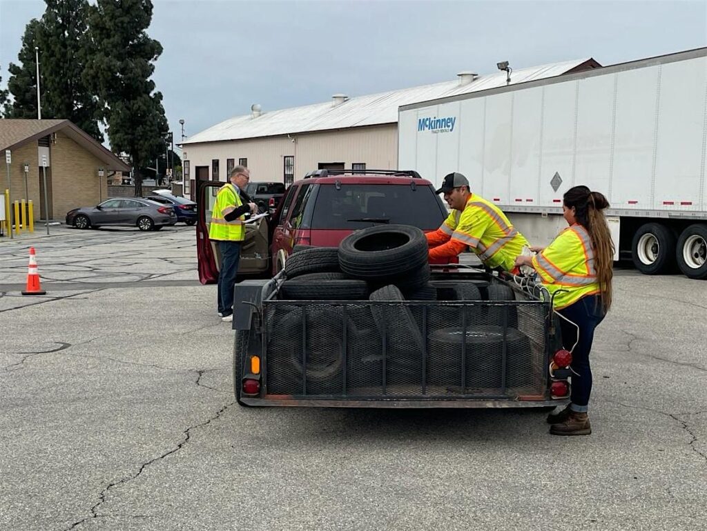 Three staff members assist in unloading used car tires from a trailer at a tire recycling event.