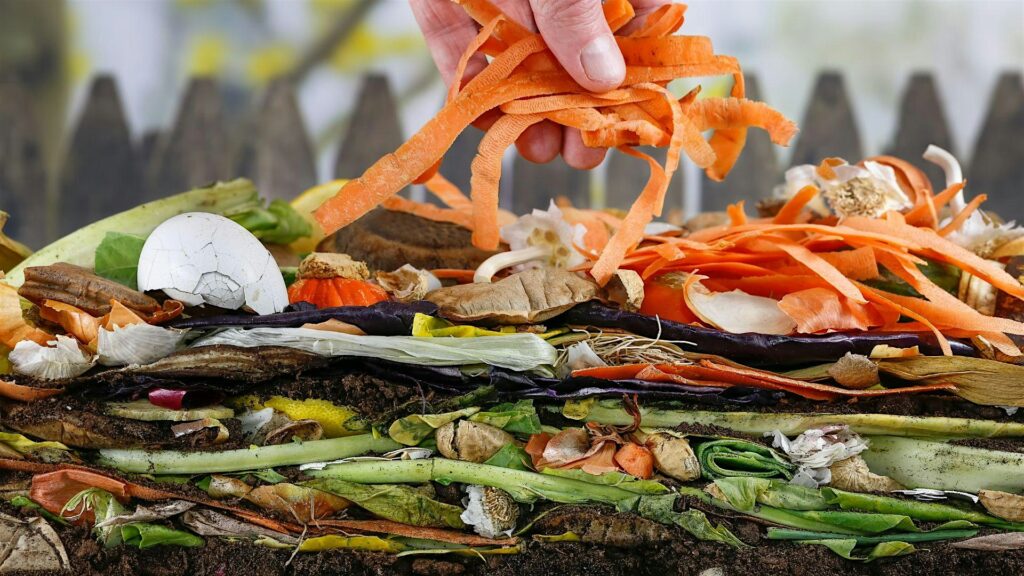 A person's hand is seen adding carrot skins to a layered pile of organic waste for composting.