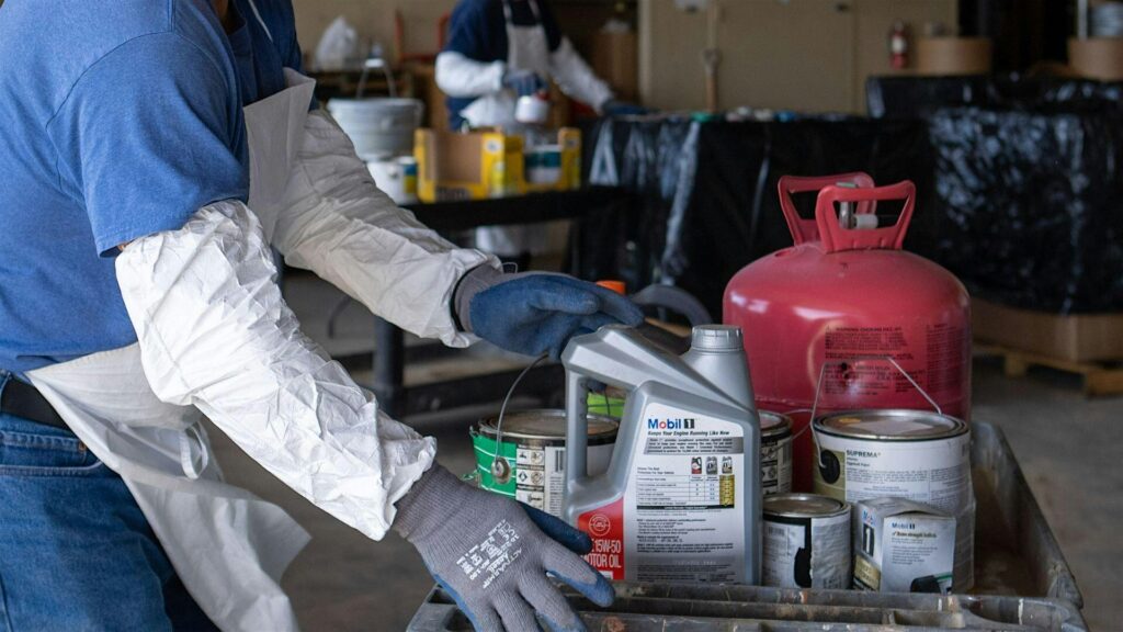 A person in protective clothing and gloves in shown collecting household hazardous waste including used motor oil, a propane canister, and house paint.