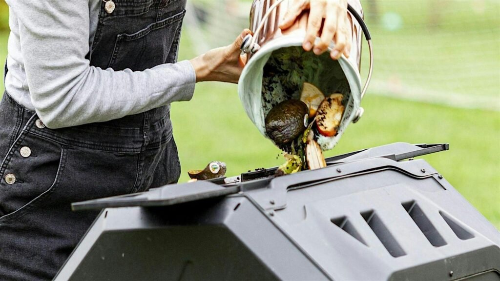 A woman in overalls empties a countertop food scrap container into a composting bin.